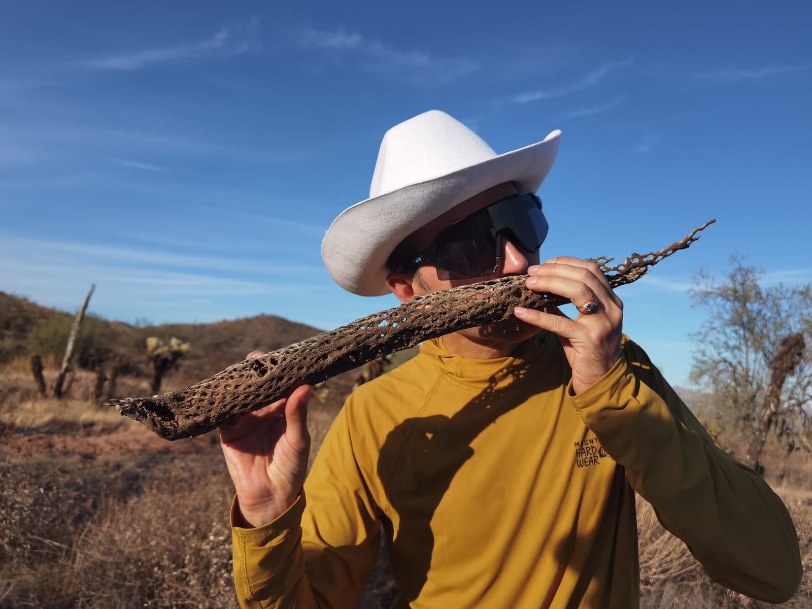 Eli in a white cowboy hat and sunglasses holds a dried cholla cactus skeleton to his lips like a flute, in the Arizona desert